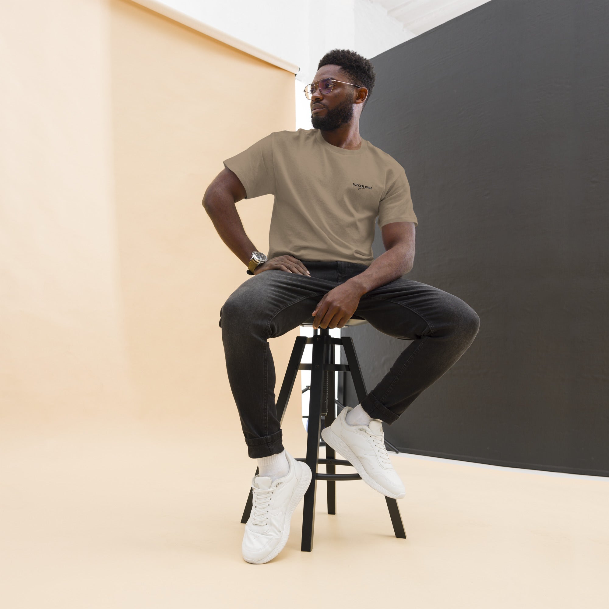 Man sitting on a stool wearing a SUCCESS WIRE Brown Savana color t-shirt with SUCCESS WIRE logo, black pants, and white sneakers against a plain background.